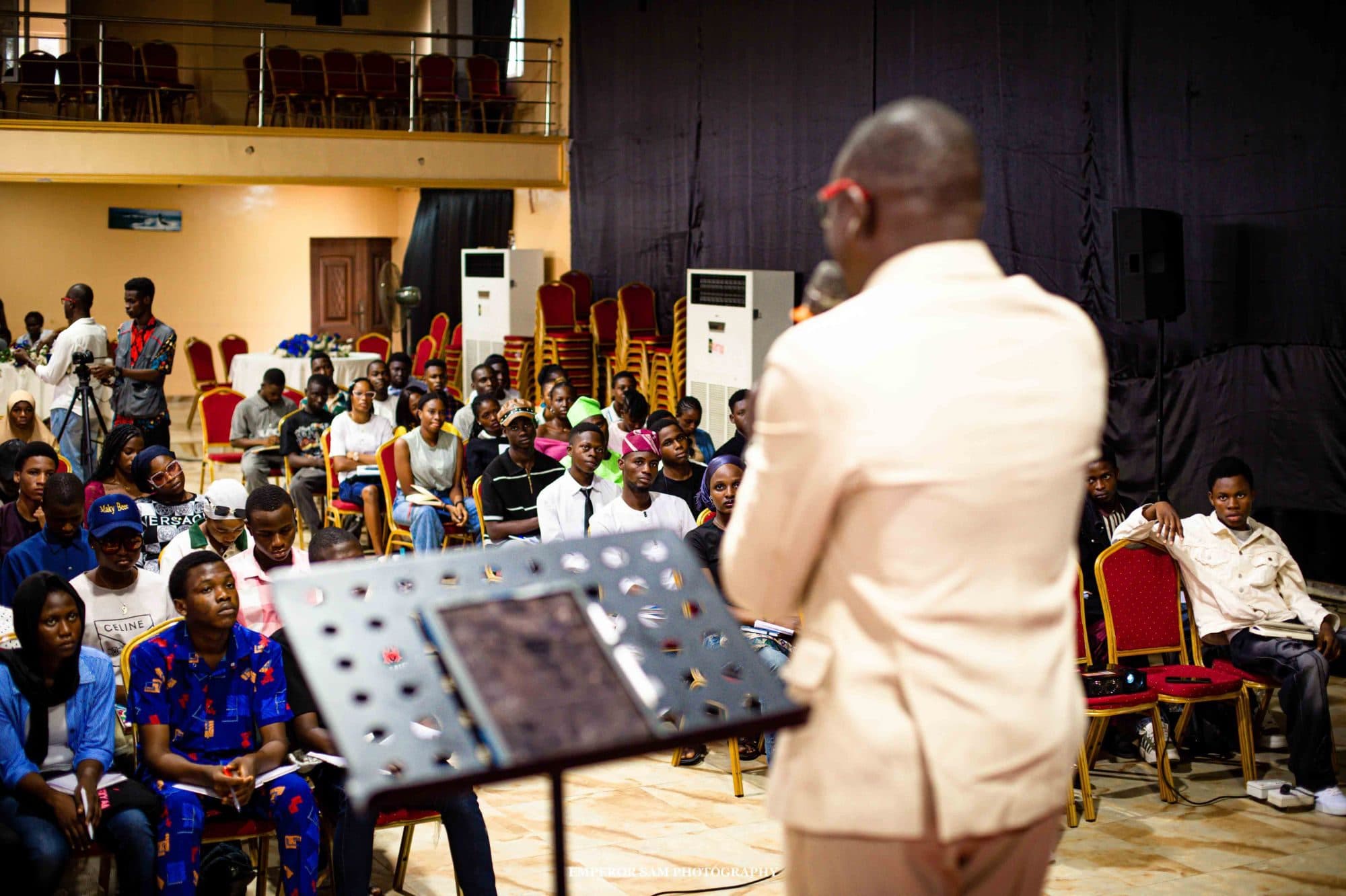 A speaker in a cream suit addresses a seated audience taking notes